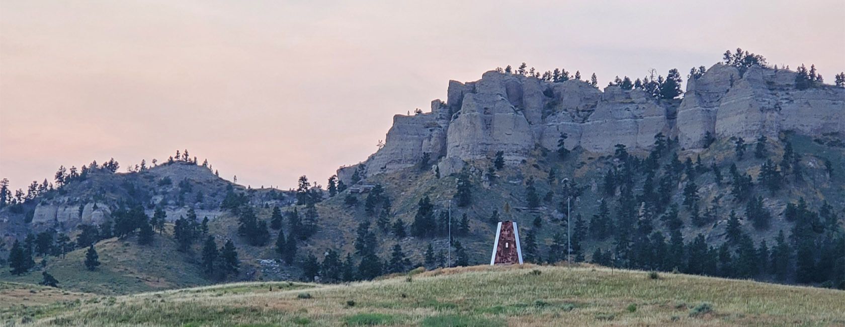 Rocky, tree-covered hill under a cloudy sky, with sparse snow patches on a grassy field in the foreground.