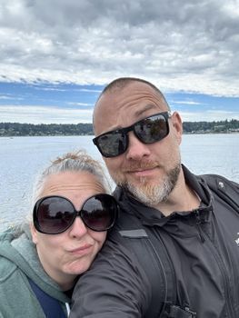 L- White woman grey hair pulled back, large sunglasses, rests her head on Rick's shoulder. Rick, (R), greying beard and mustache, close shaved head. A body of water is behind them. They are making funny faces into the camera.