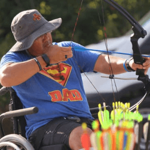 Man in a wheelchair wearing a blue t-shirt pulling back to shoot an arrow at target