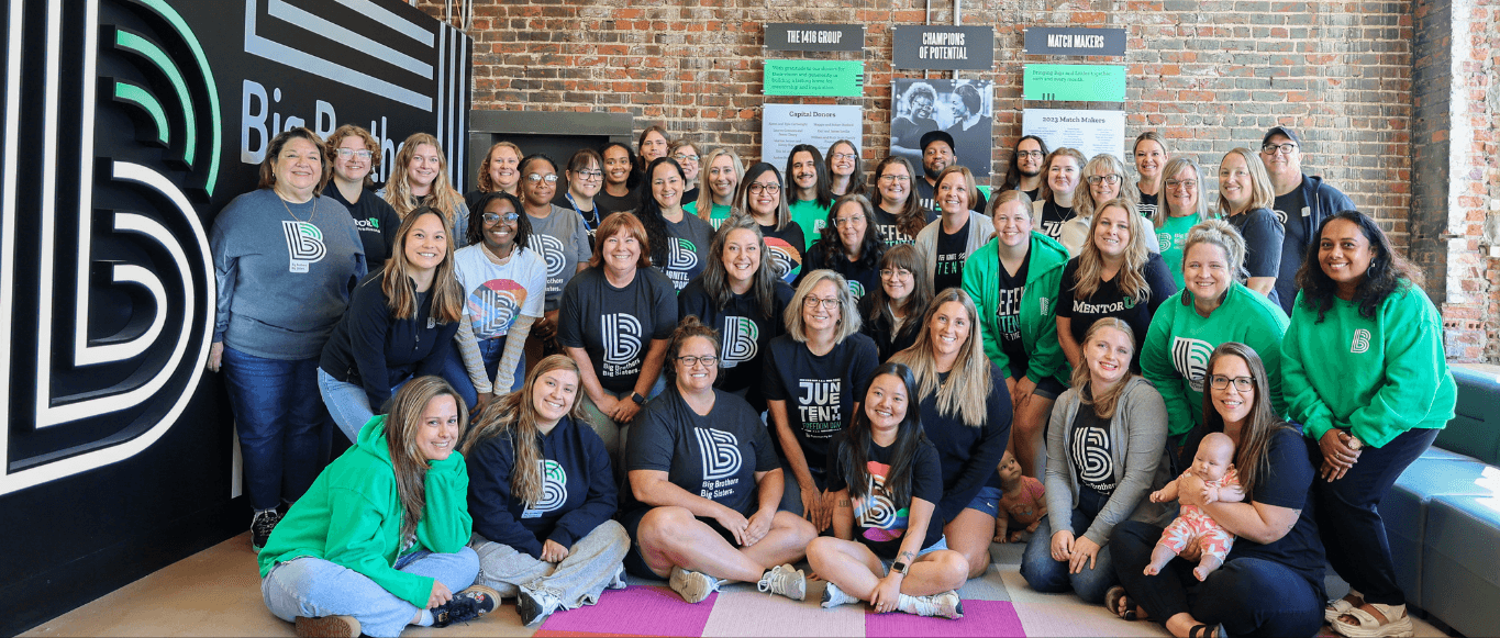 Group photo of Big Brothers Big Sisters of the Midlands staff gathered inside their office, smiling together in front of a large BBBS logo wall. Team members wear organization-branded shirts and sweatshirts in green, black, and gray, representing unity an