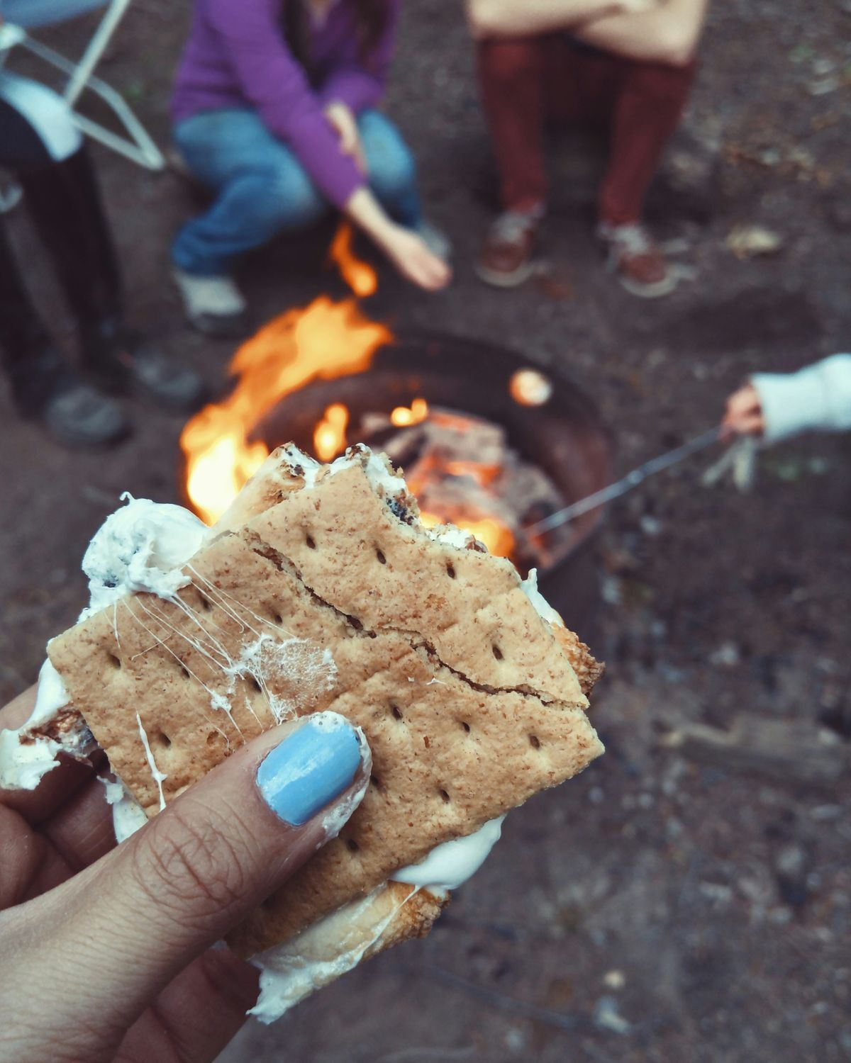 A partially eaten s'more backed by people roasting marshmallows over a fire