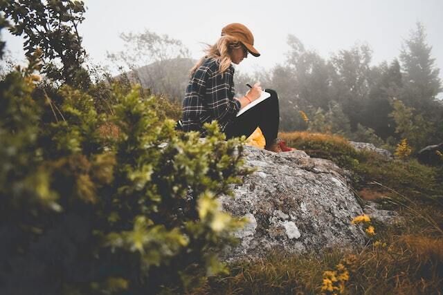 Blonde haired white woman wearing a hat sitting on a boulder and writing in a notepad