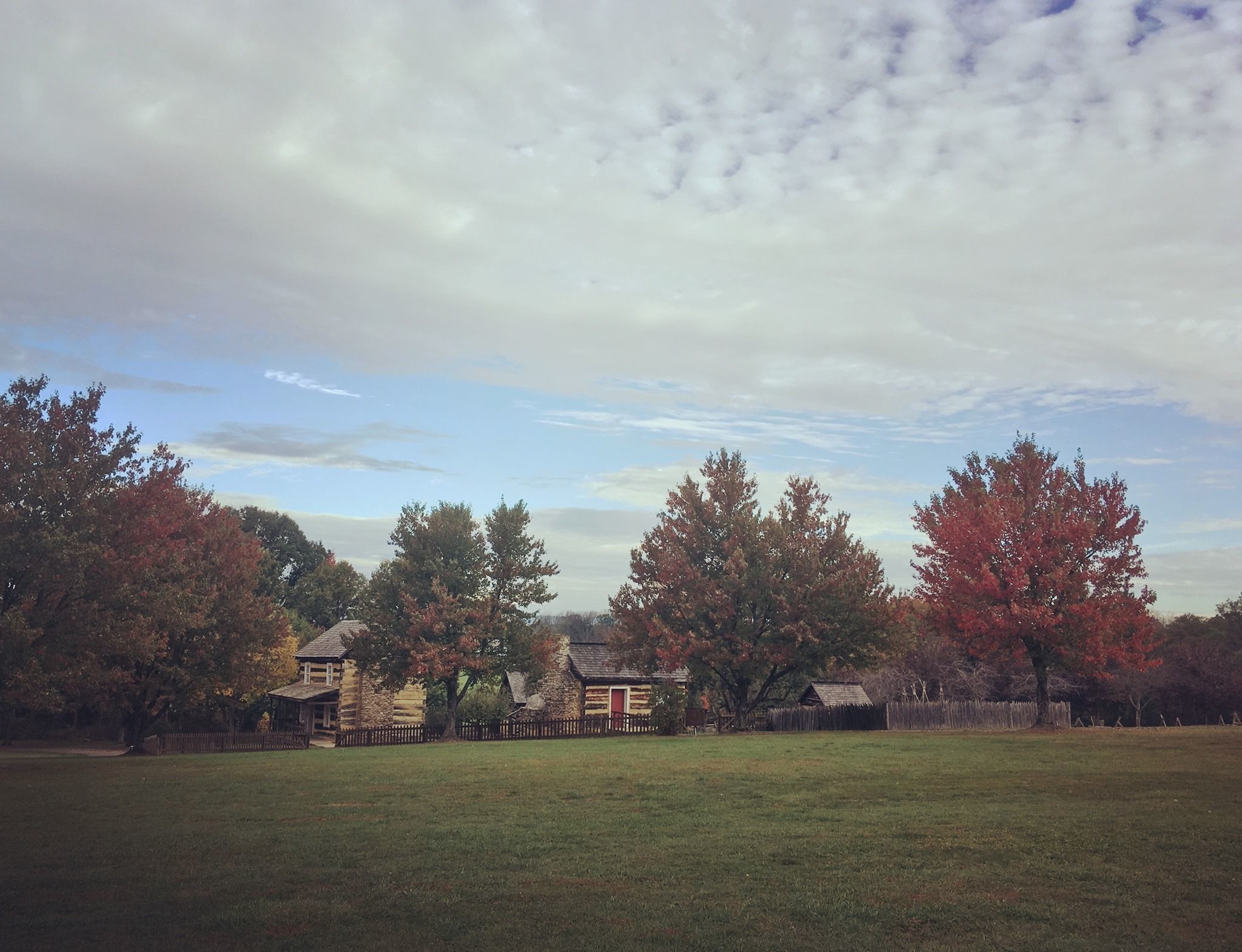 Picture of a log farmstead in spring with new leaves and a partly cloudy sky.