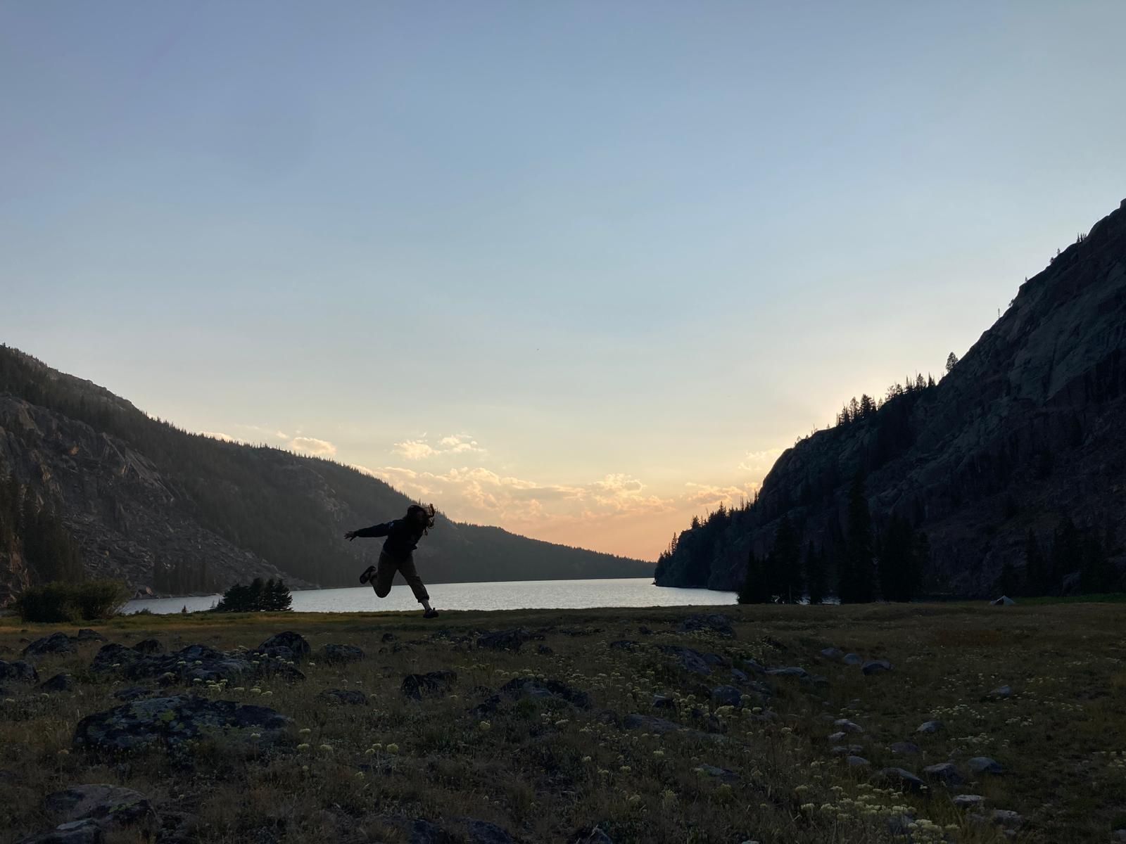 A crew member jumps up into the air. In the background is a lake with the sun setting behind it.