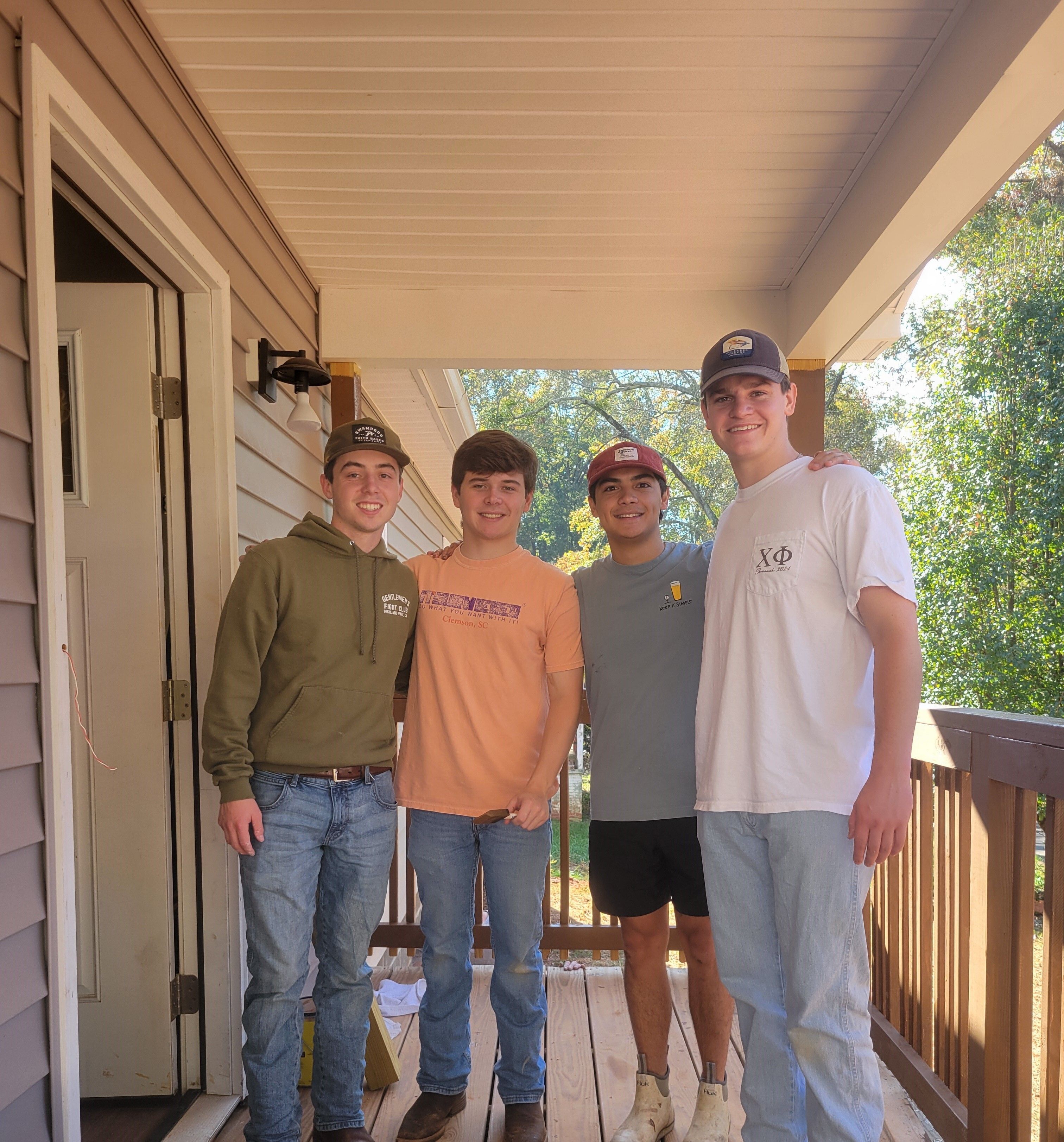 Four Chi Phi fraternity members proudly standing on the porch of a house they helped bring to life, smiling and celebrating their community service efforts. The image captures their camaraderie and commitment to giving back.