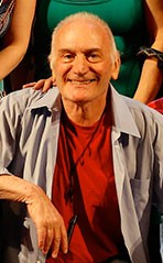 IKE SCHAMBELAN, A bald man with a big smile on his face. He is wearing a red t-shirt and a gray shirt over it. His arm is leaning on something to his right. He is facing the camera directly.