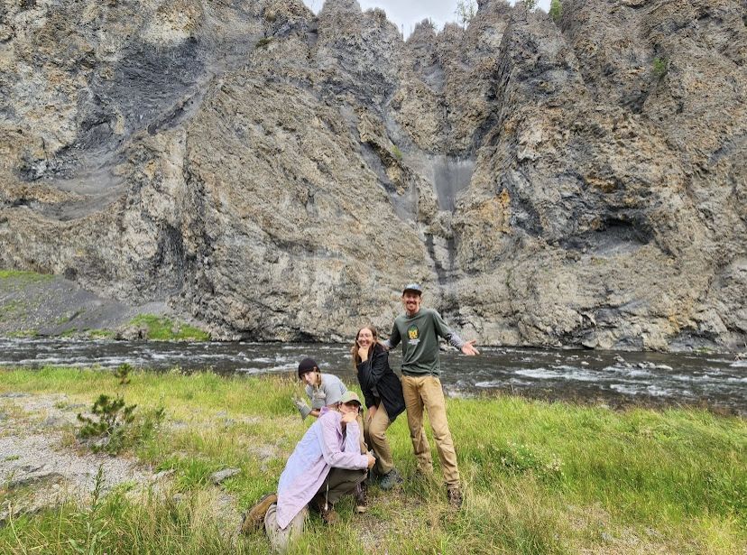 A crew makes goofy poses in front of a rock face, with a small stream in the background.