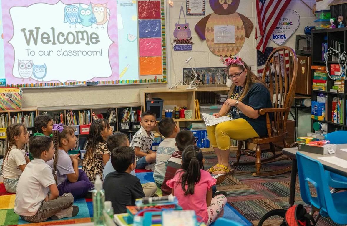 A teacher sits in a rocking chair reading to a group of young students gathered on the carpet, with a colorful “Welcome to our classroom” sign displayed on the wall.