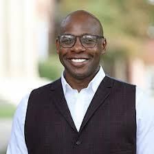 Dr. Reginald Ball stands outdoors, smiling warmly at the camera. He is wearing glasses, a white shirt, and a dark vest, with a blurred background of greenery and buildings behind him.