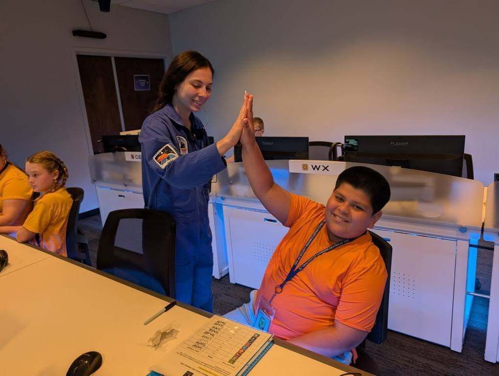Commander Mya high-fiving student in orange shirt while in our mission simulator. 