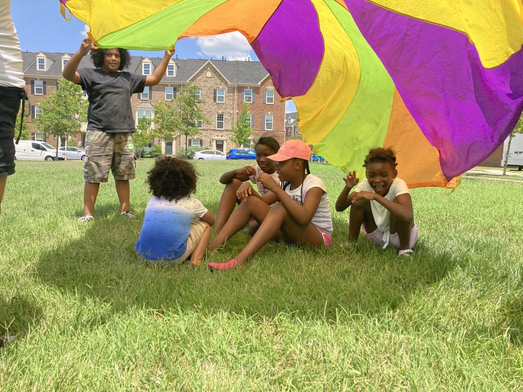 Children on a grass field sitting under a rainbow parachute while another child lifts it over their heads.
