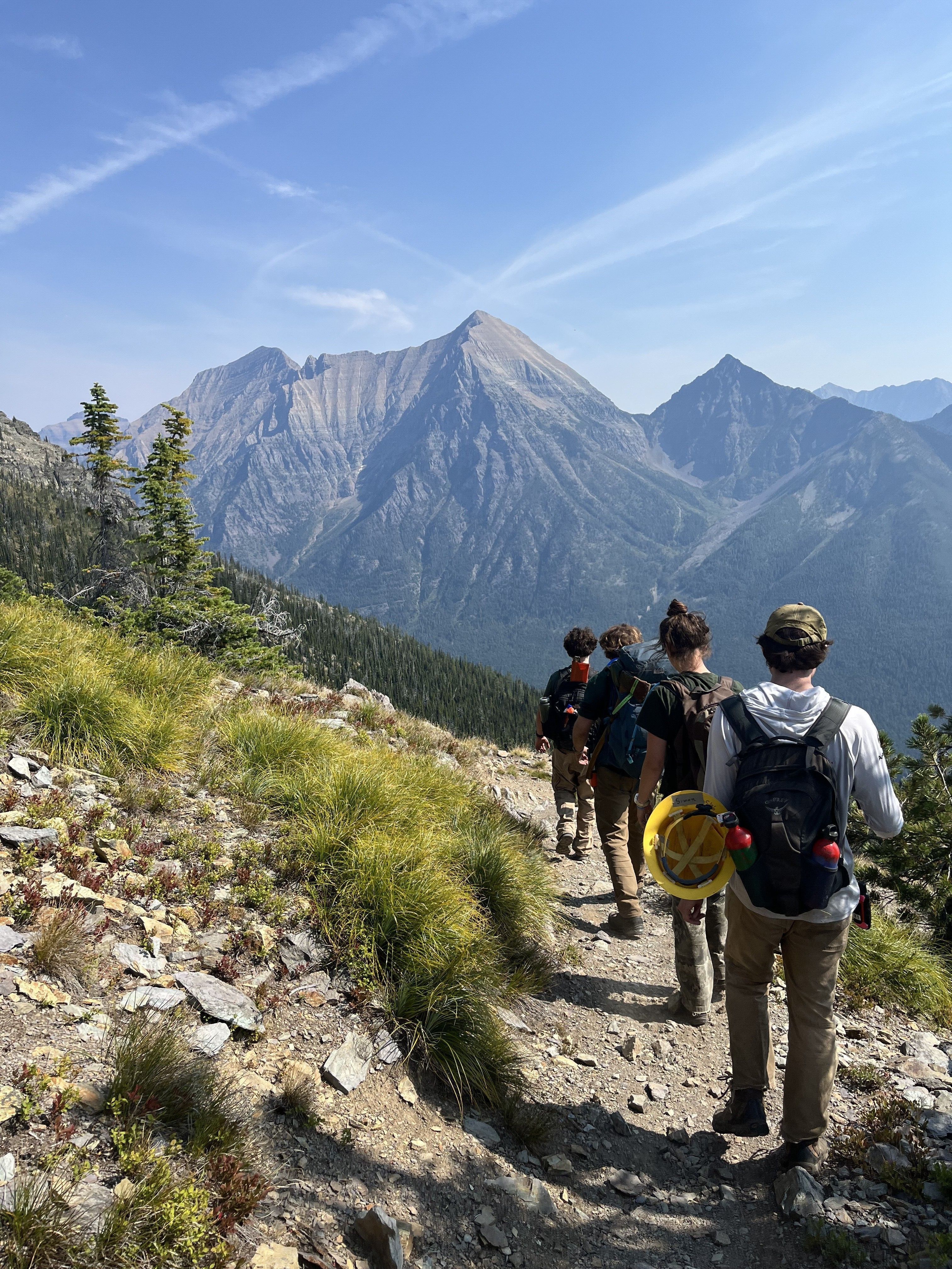 [Image Description: Four MCC members are walking away from the camera, along a trail with large mountains in the distance.]