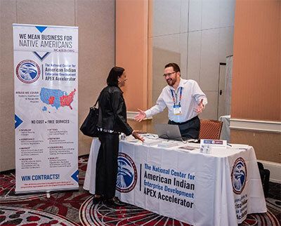 Two people converse at a booth for the National Center for American Indian Enterprise Development APEX Accelerator.