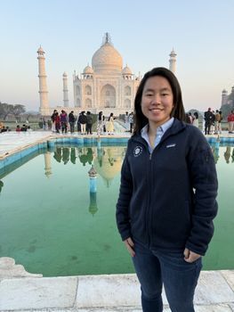 Naomi - Asian woman with black hair, smiles while standing in front of the famous Taj Mahal. She is wearing a dark jacket with a blue collared shirt underneath. Behind her, the famous white marble monument, Taj Mahal, its minarets, and a reflecting pool