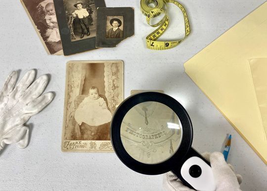 five historic photographs with one being examined with a magnifying glass. White gloves, tape measure, and archival folder also laying near by.