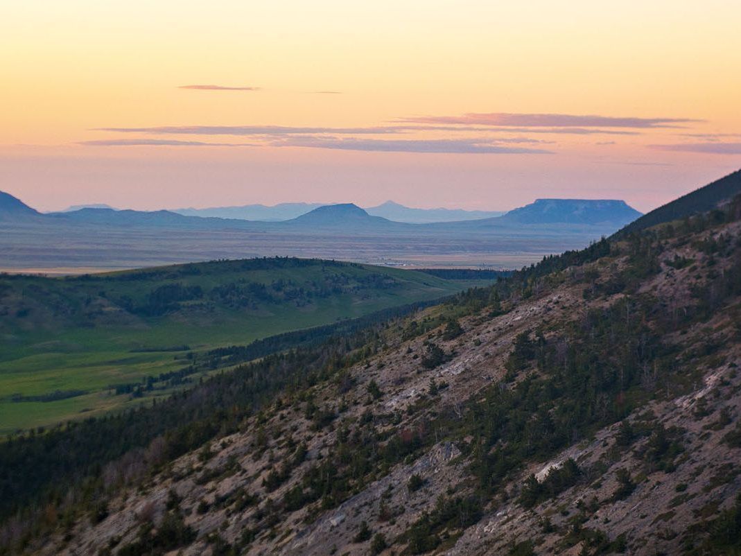 [Image Description: A panoramic view of the buttes and hills outside of the city of Helena. The sun is gently setting, coloring the sky with pastel hues of pink and yellow.]