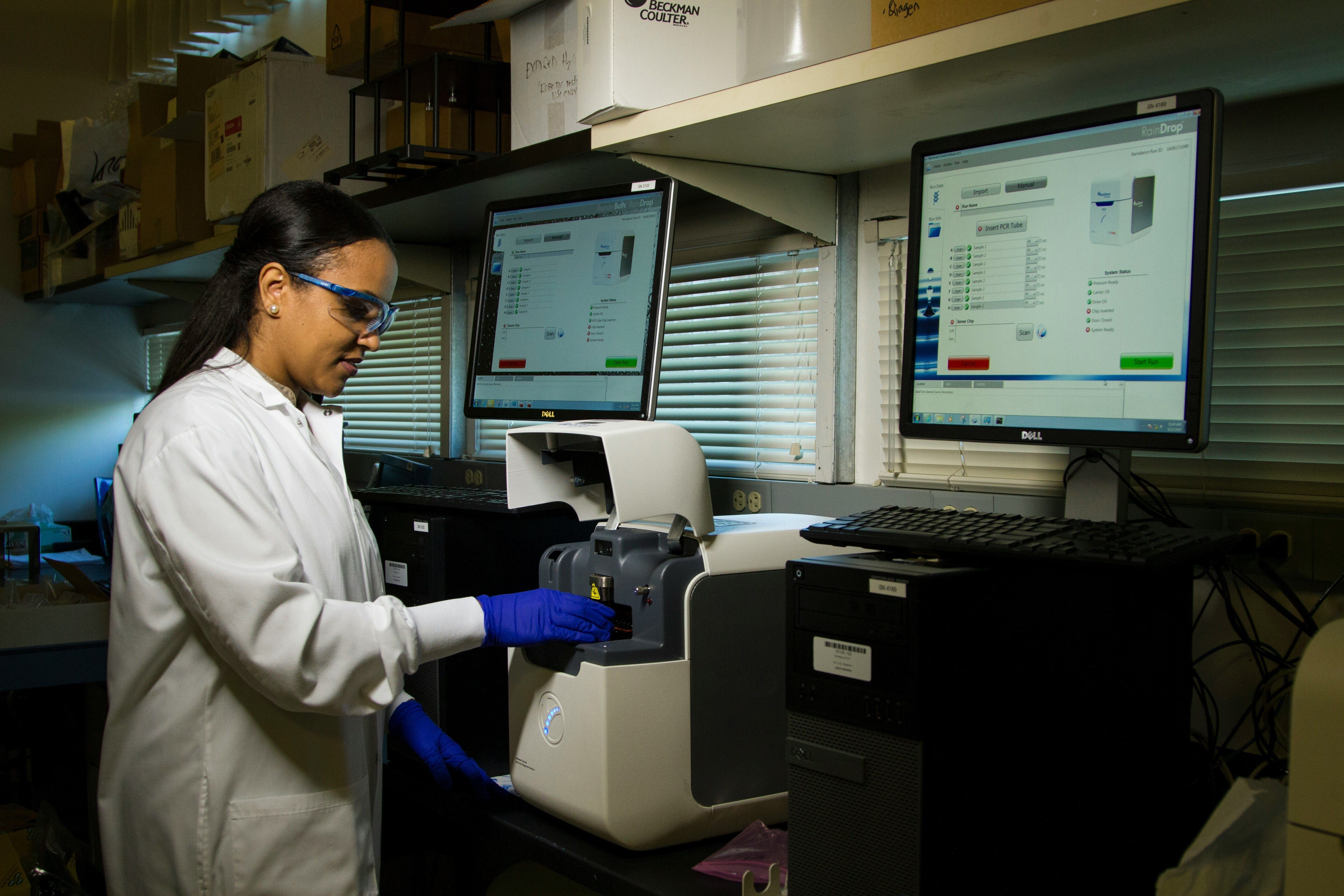 Stock image of a woman in a lab coat and blue gloves and safety classes is working on a laboratory equipment with computer monitors around her in a lab setting