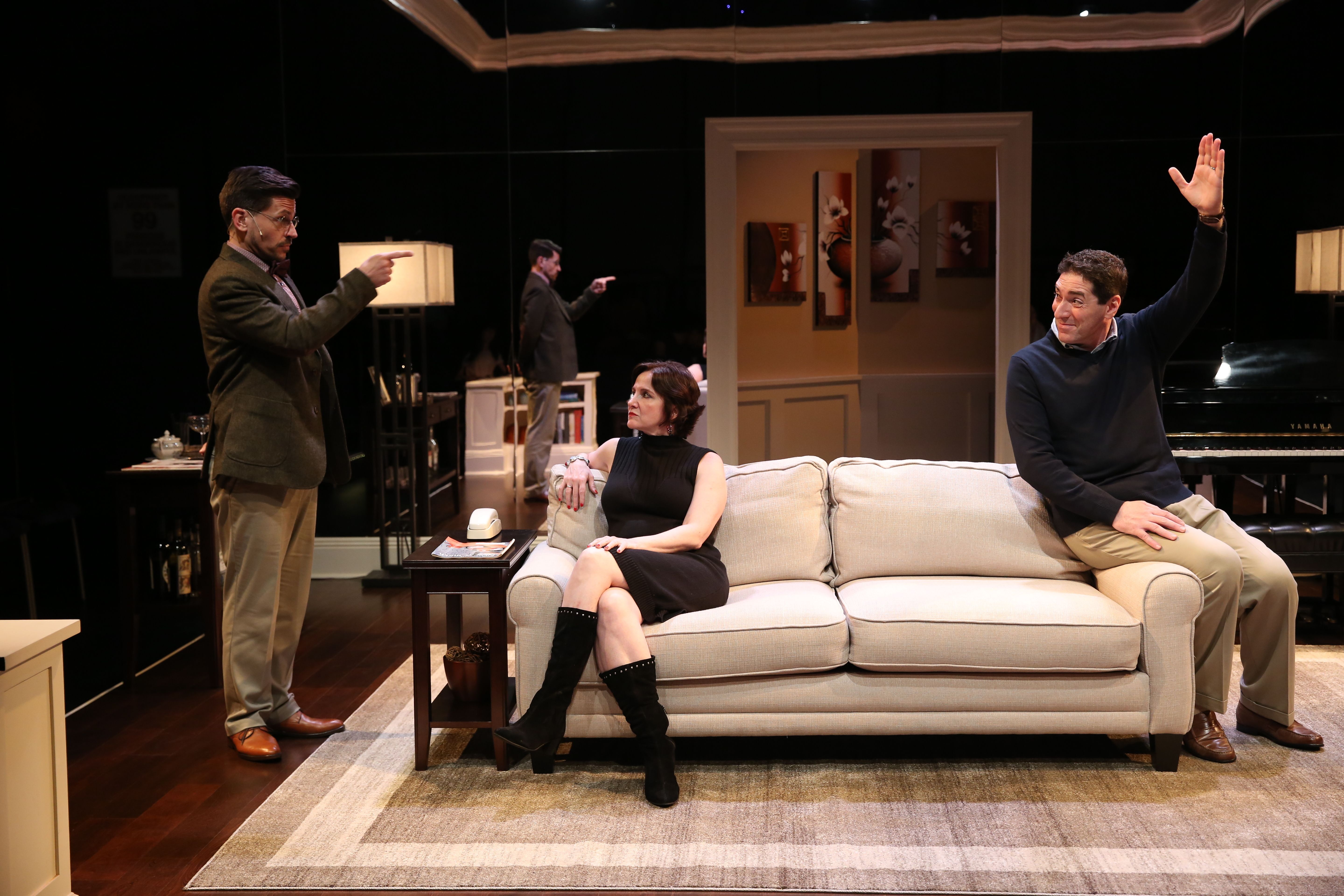 A picture of three actors who’re both sitting and standing. They’re performing a scene from the production of The Fourth Wall. They’re in a living room, and an actor on the left is pointing his finger at the actor on the right who has his hand raised. 