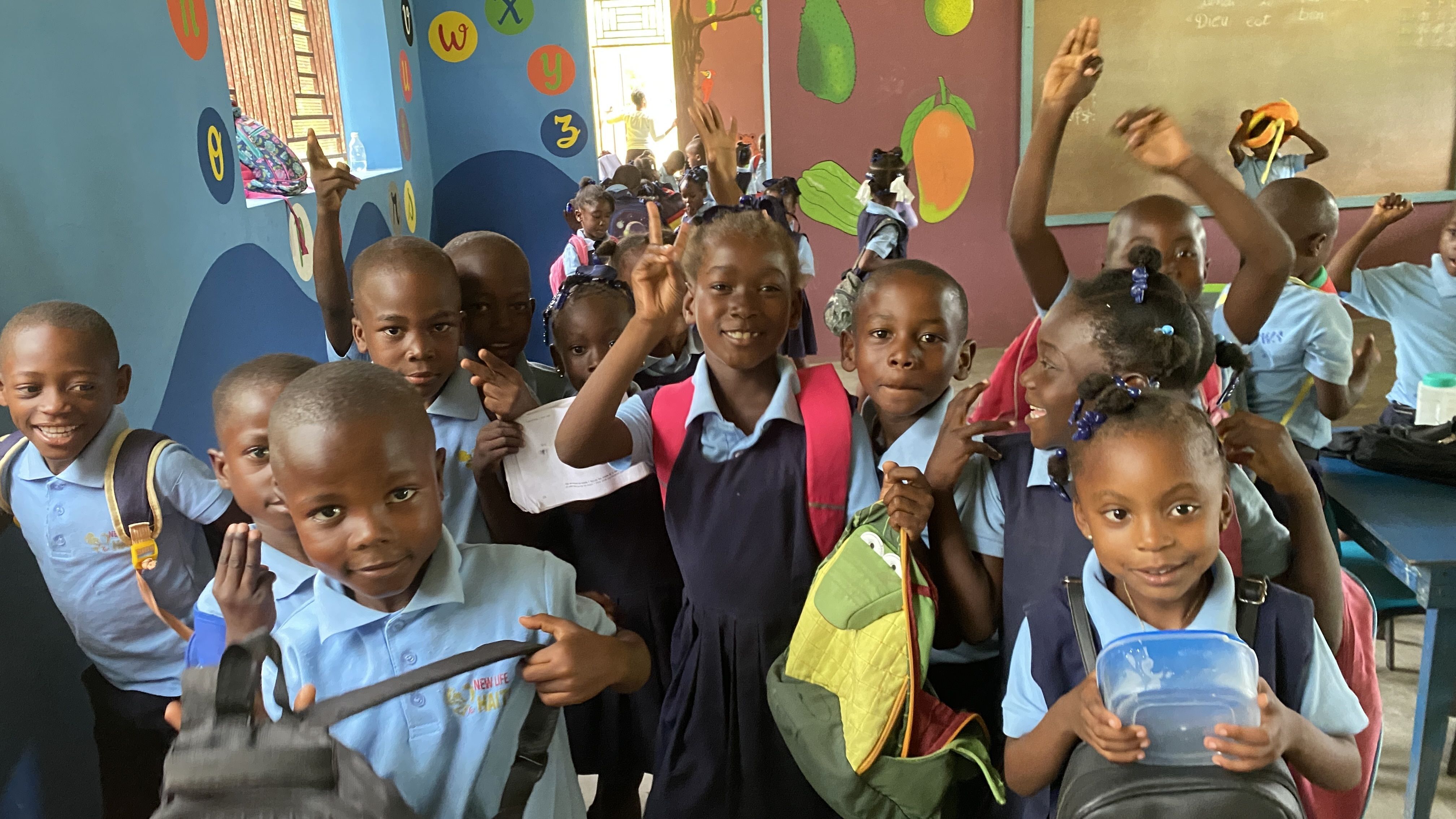 schoolchildren wearing blue uniforms and backpacks grin widely in their colorful classroom