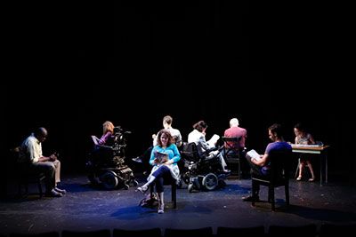 A dark scene with people who are both in wheelchairs and in regular chairs. They are all seating in in a circle and reading articles on different platforms, but they are all facing outward. One person is reading at the table in right corner of the room. 
