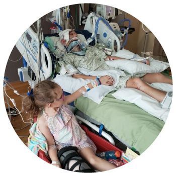 Carson lying in a hospital bed after the accident. Lilly sitting next to Carson holding his hand.