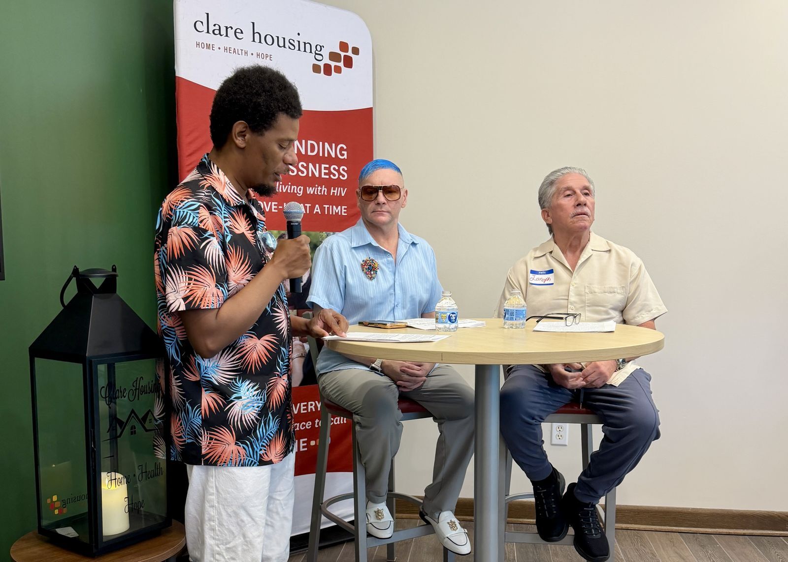 A person stands with a microphone and two people sit at a high-top table