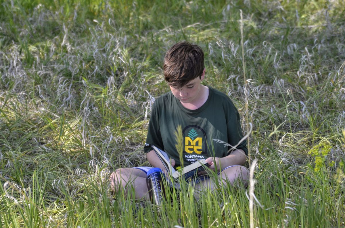 [Image Description: An MCC youth crew member is sitting in a field, reading their curriculum in a grassy field.]