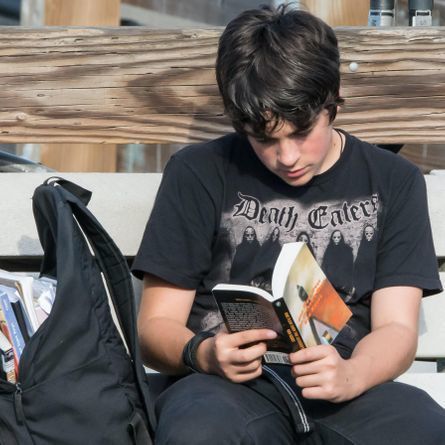 Teen with a backpack full of homework reading on a bench outside.