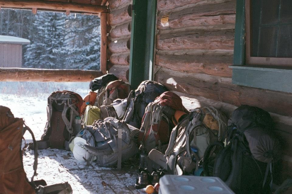 [Image Description: Nine fully packed backpacking backpacks are resting on the outside of a log cabin. There is a dusting of snow on the ground and on the porch of the cabin.]