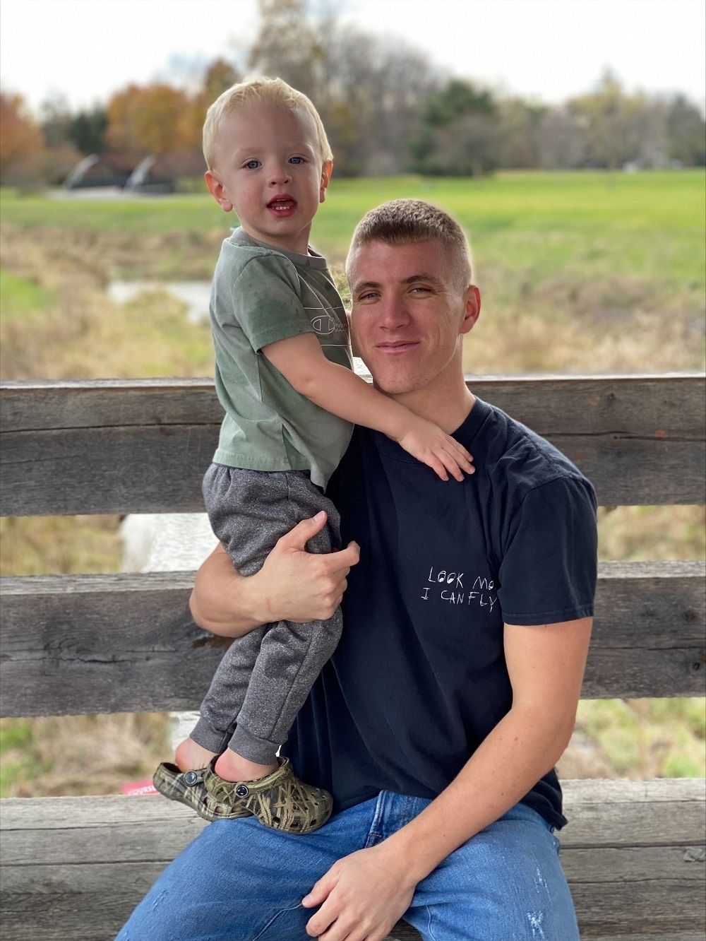 Maxwell holds his young son while sitting on a wooden bridge on a nice day.