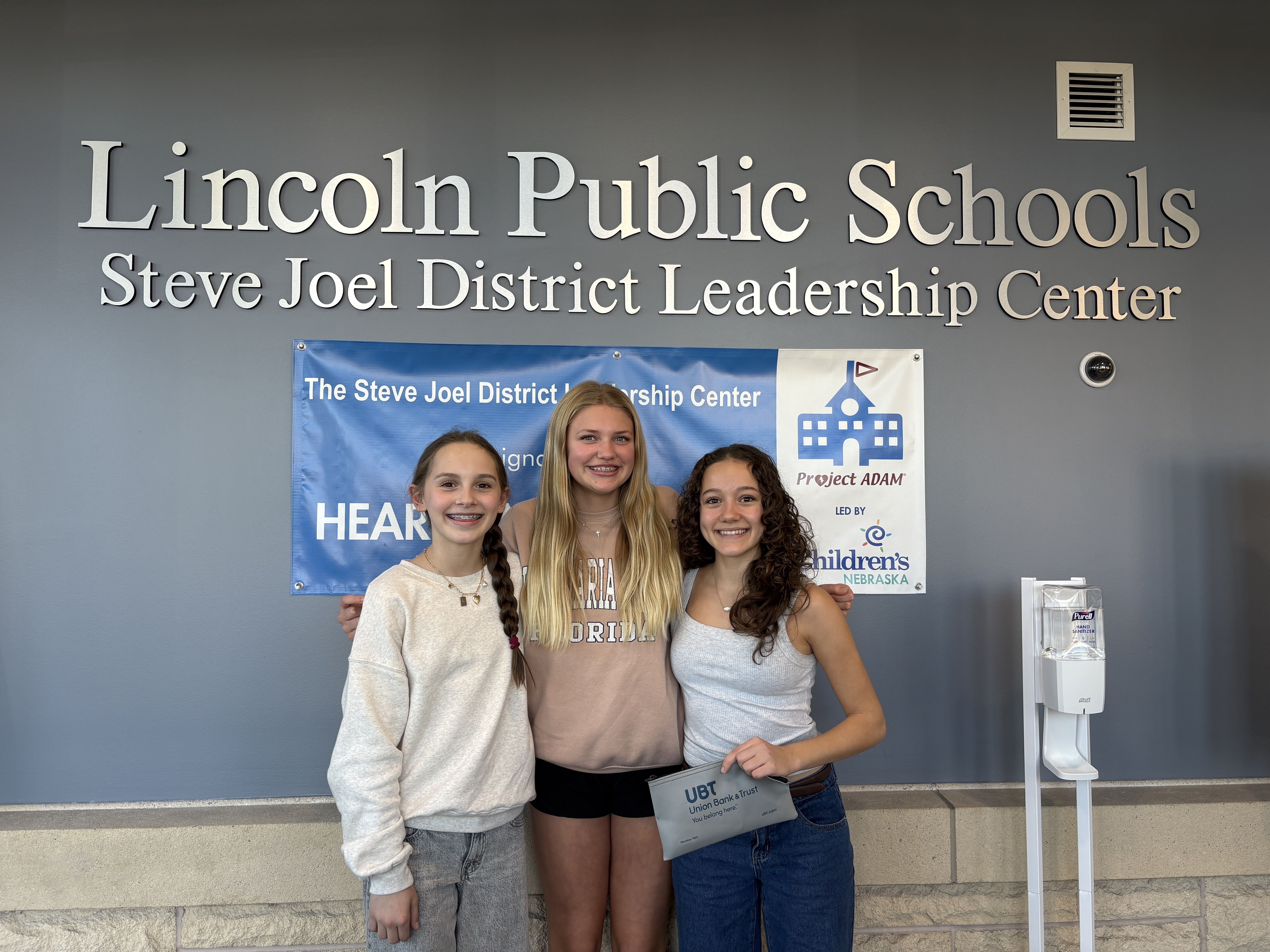 group of three middle school girls posing with check from their bake sale in front of LPS sign