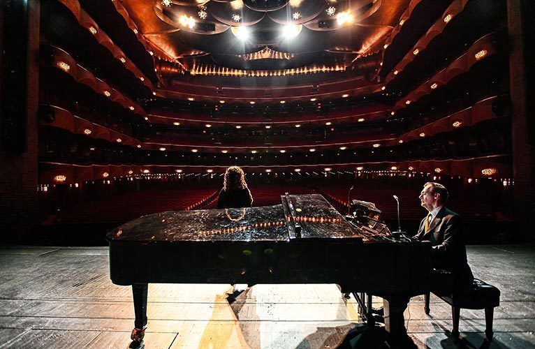 A young singer with curly hair stands in front of a piano played by a middle aged gentlemen in a gray suit. She is facing the empty seats of the Metropolitan Opera House in New York City.