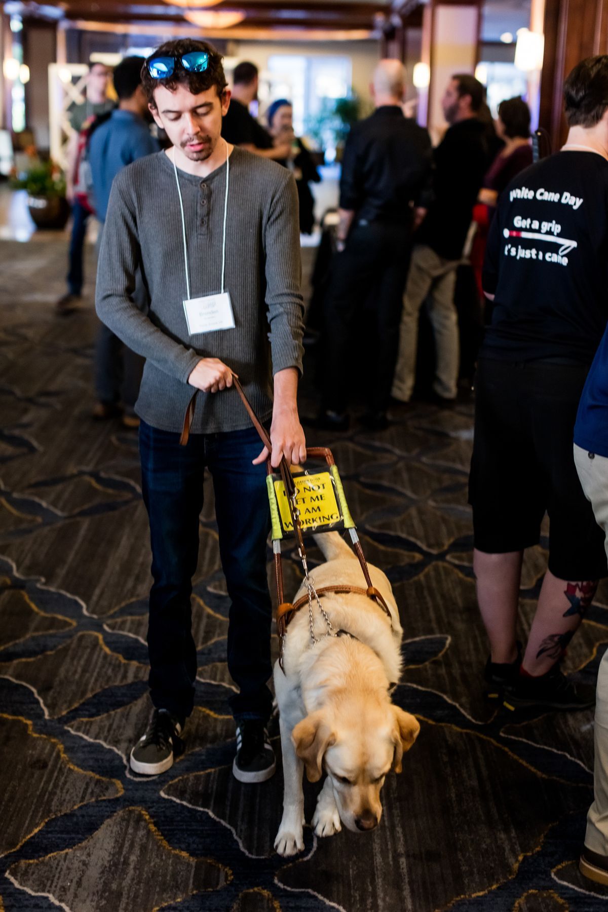 A young man with brown hair, wearing a grey shirt and black pants is holding his guide dog's harness. His guide dog is a yellow golden retriever. 