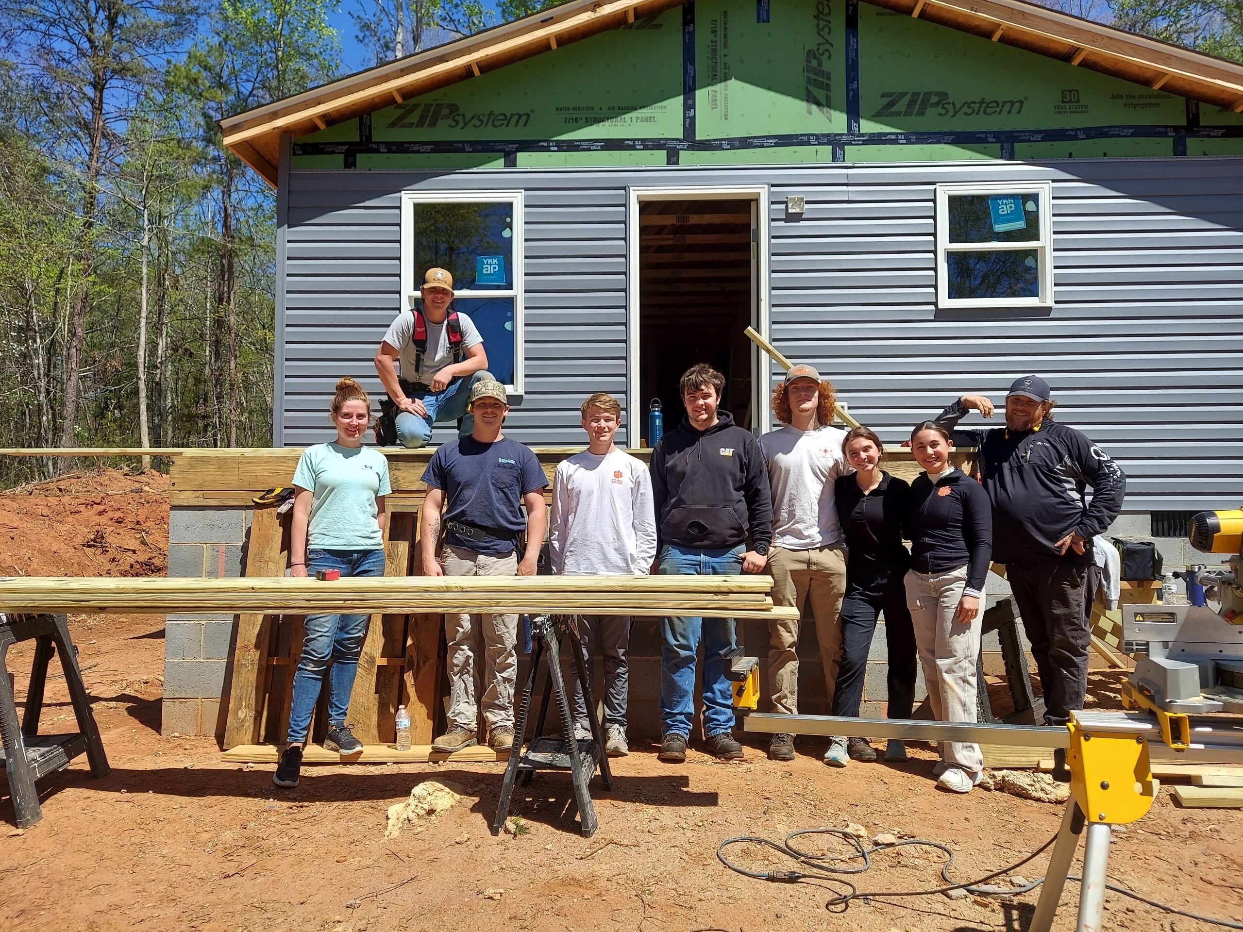 Volunteers pose in front of a Habitat for Humanity home under construction in Pickens County, SC, on a sunny spring day.