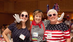 Three women stand side-by-side. They are all wearing red, white, and blue costumes. The one in the middle has her arms around the other two.