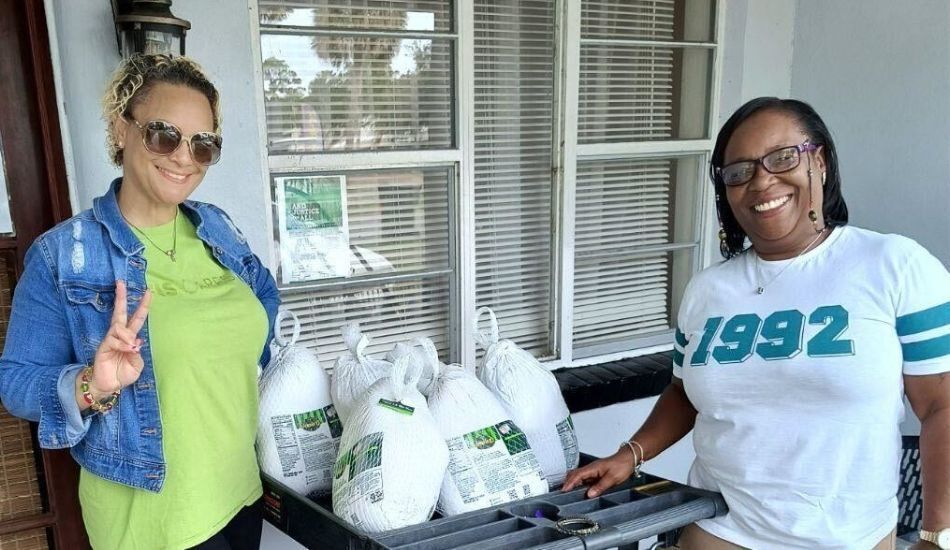 Photo of two smiling volunteers at the food pantry, with a cart full of frozen turkeys for the holidays. One wears a bright green tshirt with a jean jacket, the other wears a white tshirt with "1992" in teal letters across the chest.