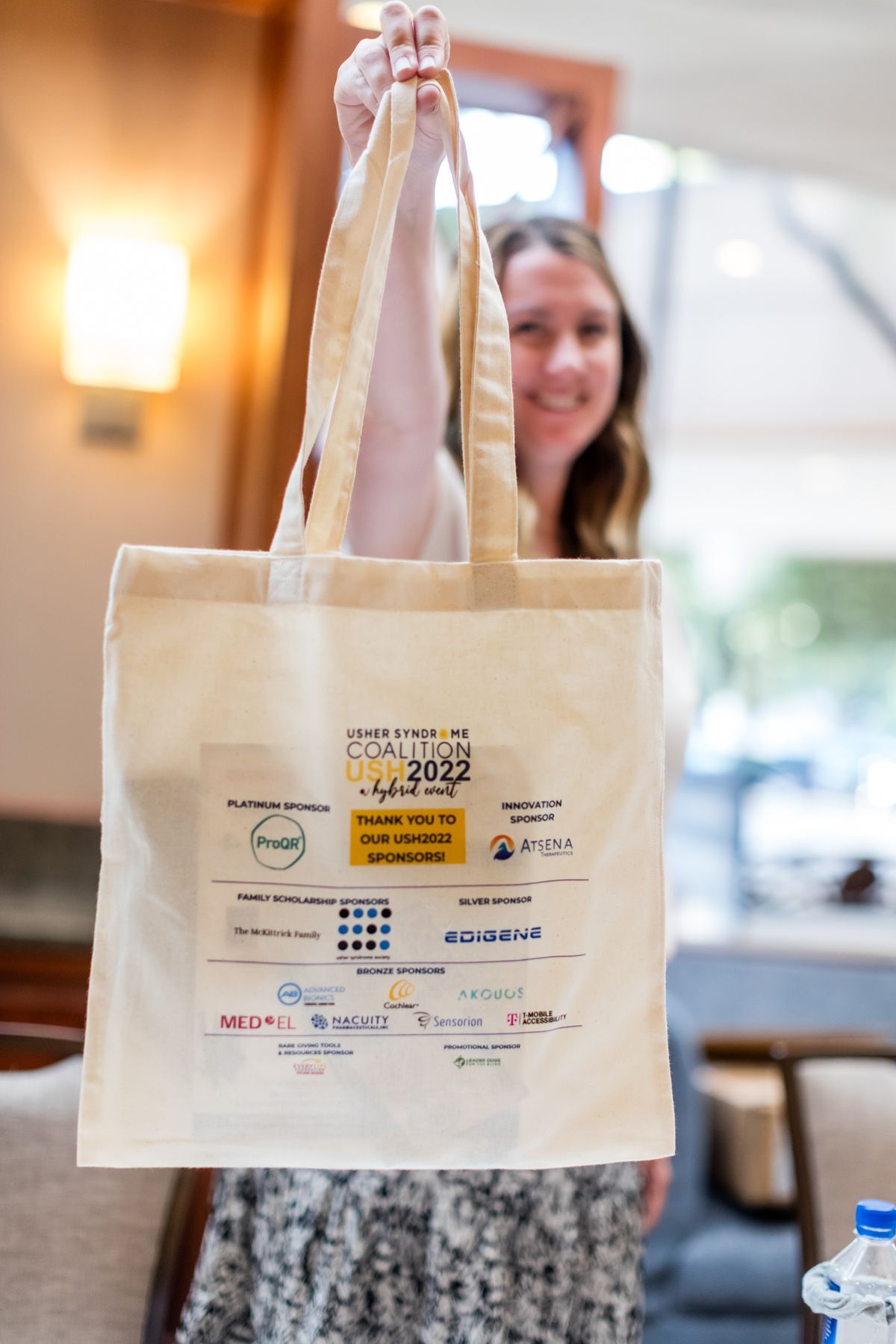 A woman is holding up the USH 2022 Connections Conference Tote Bag that is a light tan color with all of the Conference sponsors logos on the bag.