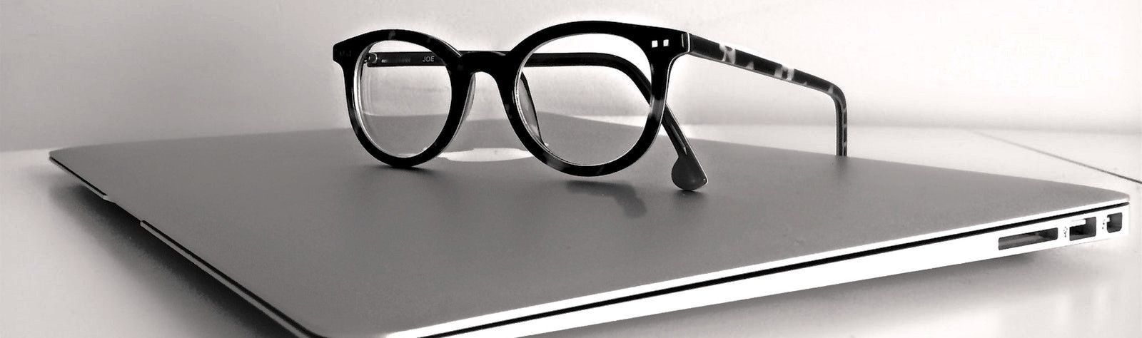 Sideview photo of a pair of round, black horn rim glasses sitting on top of a closed silver Mac laptop. The laptop is set on a white table and the background is light grey.
