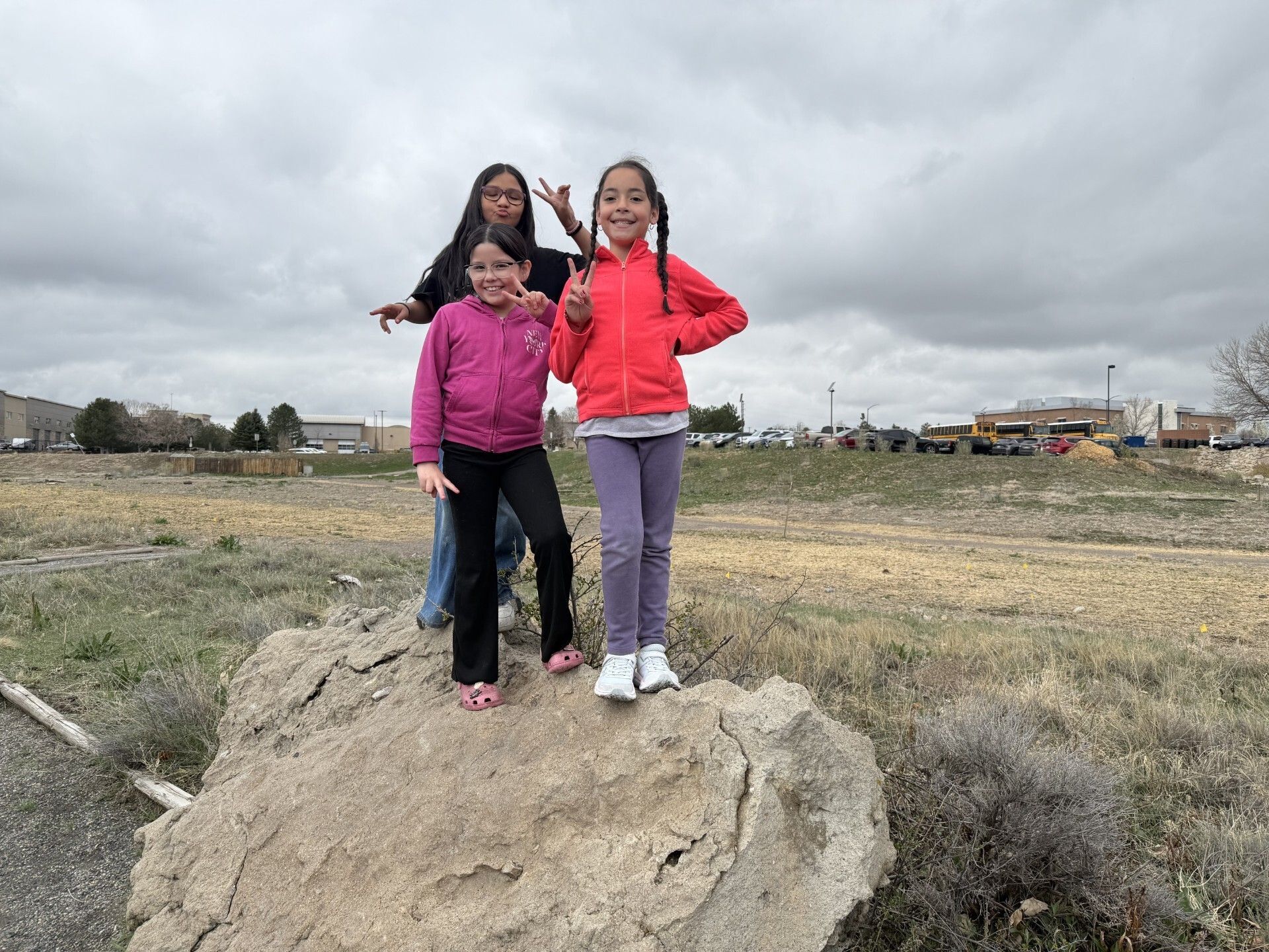 Three smiling children stand on a large rock in an open field near a school, symbolizing strength, joy, and the uplifting spirit of MEF school-based Chapters.
