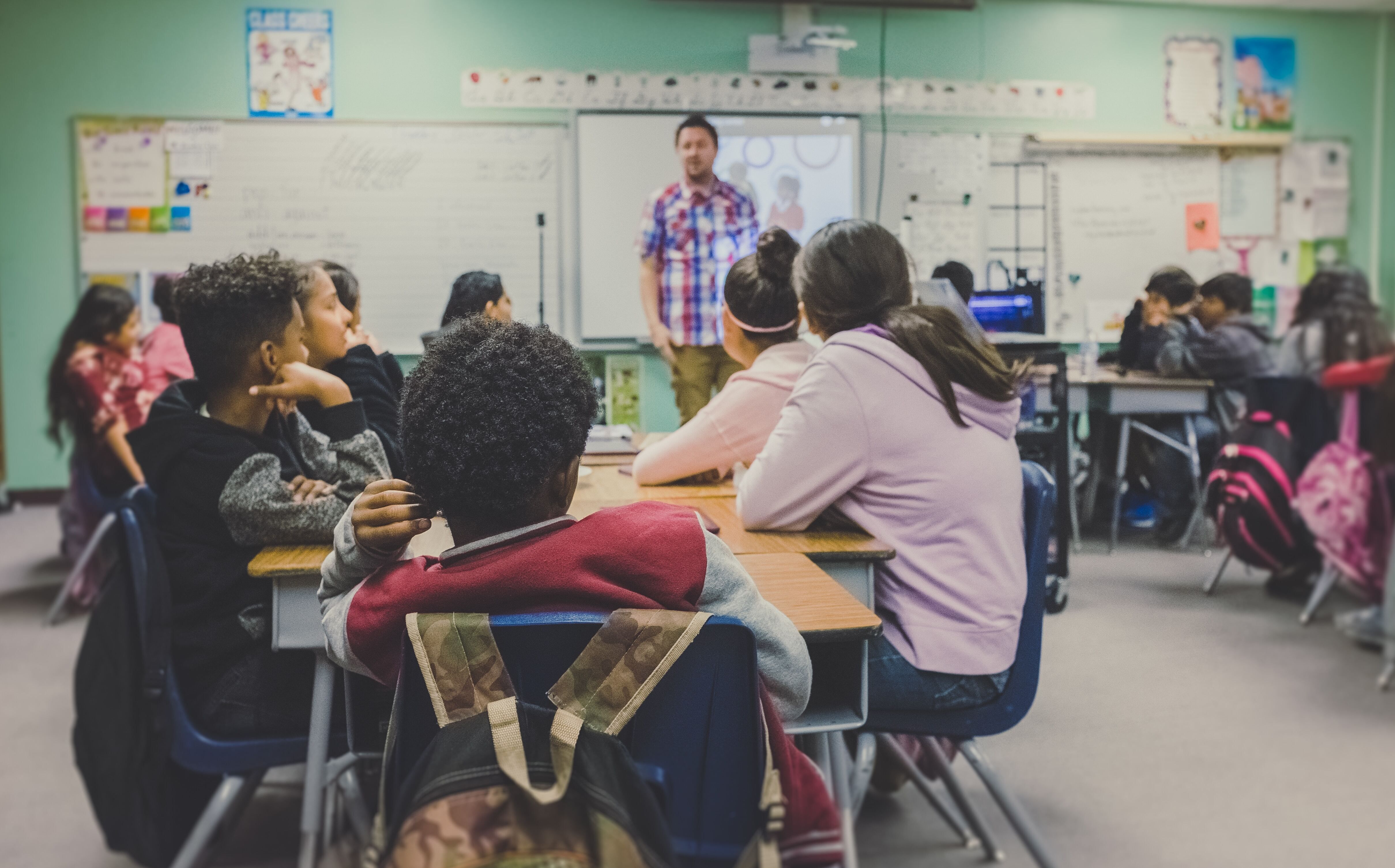 Male teacher standing in the front of a classroom. Middle school age kids sitting in the classroom.