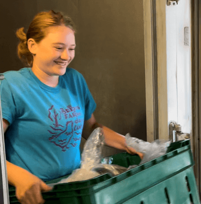 A teenage girl in a light blue t-shirt is carrying a plastic crate of produce from left to right across the photo