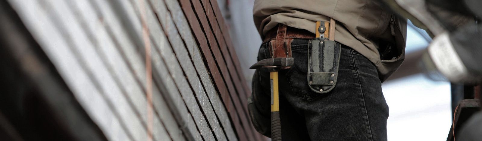 Sideview photo of a construction worker's tool belt on his waist. He wears a tan jacket and black jeans. From his belt hangs a hammer with a yellow handle and another tool that is sheathed. He stands with his back to a wall with boards that are red-brown,
