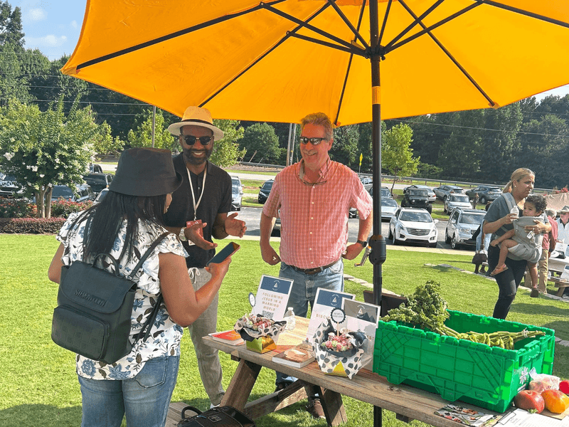 People talking at a picnic table with produce under an umbrella