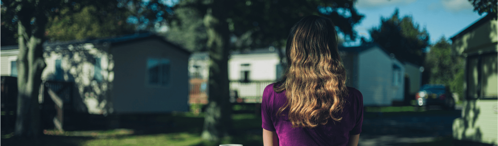 Photo of a young woman looking out on a row of manufactured houses. She is just right of center, wearing a short sleeved fuchsia t-shirt and has dark blonde wavy hair that falls several inches past her shoulders. In the background there are three manufact