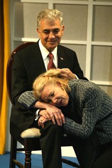 George is sitting on a chair, wearing a suit and tie. He's touching Melanie's hair who has her head in his lap. Melanie is wearing a gray pattern suit.