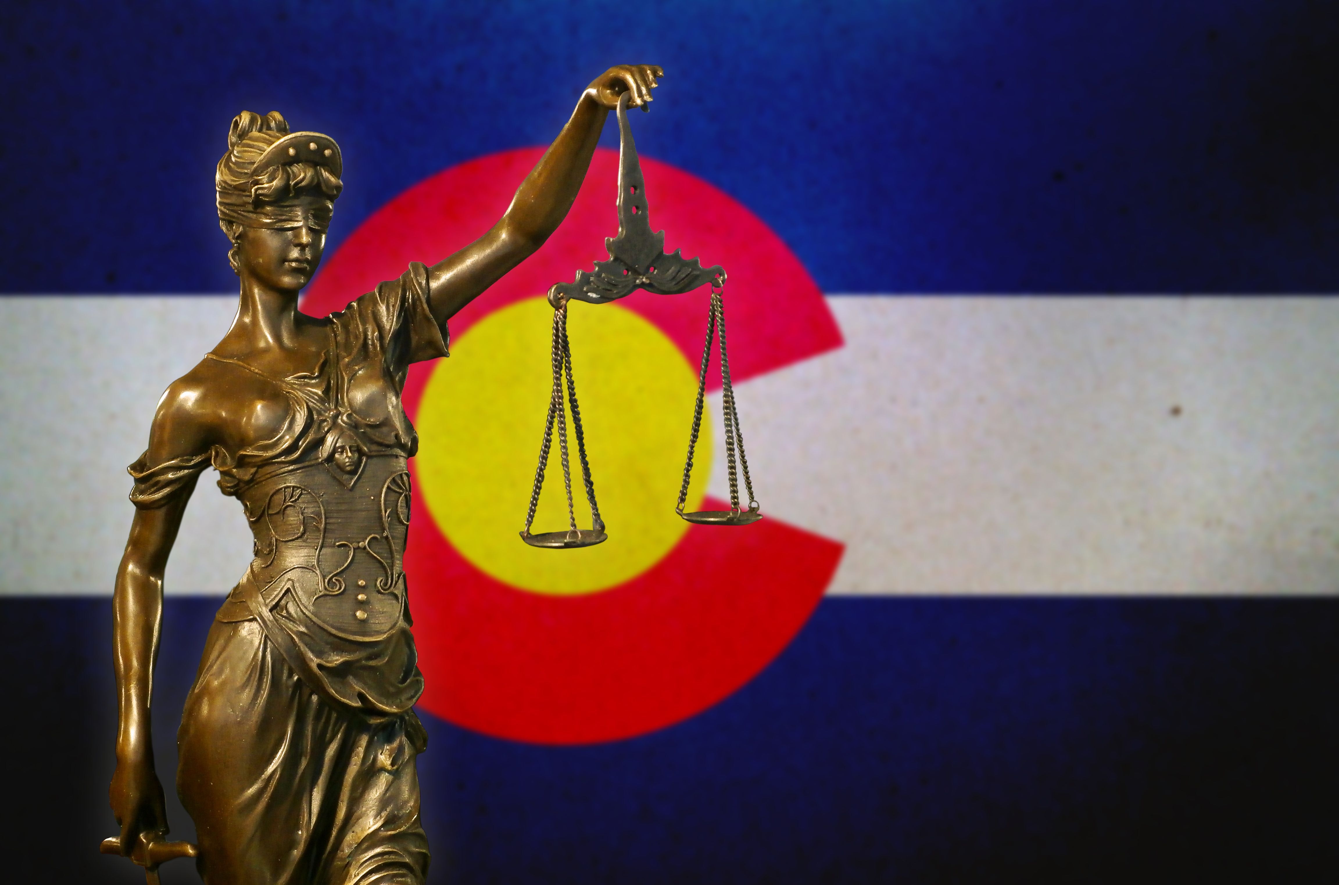 Blindfolded Lady Justice holding scales against a backdrop of the Colorado state flag